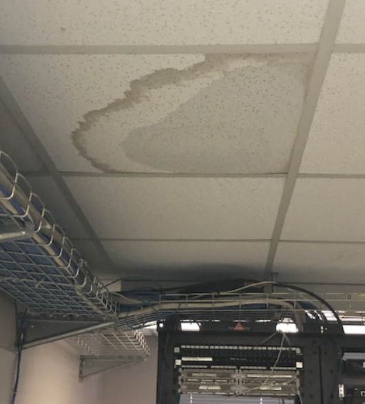 A water-damaged ceiling tile with a large brown stain hangs above computer servers and wire conduits.