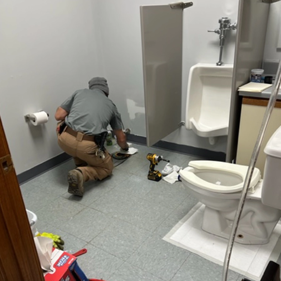A maintenance worker kneels on a bathroom floor, cleaning while surrounded by tools, paper towels, and a toilet in a simple, utilitarian space.